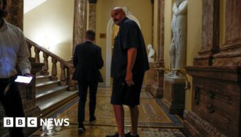 Fetterman in dark short-sleeved shirt, shorts and sneakers in a yellow hallway next to man in suit.
