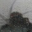 Regentropfen haften an einer Fensterscheibe eines Fahrzeugs, in der sich eine Person mit Regenschirm spiegelt. Foto: Matthias Be